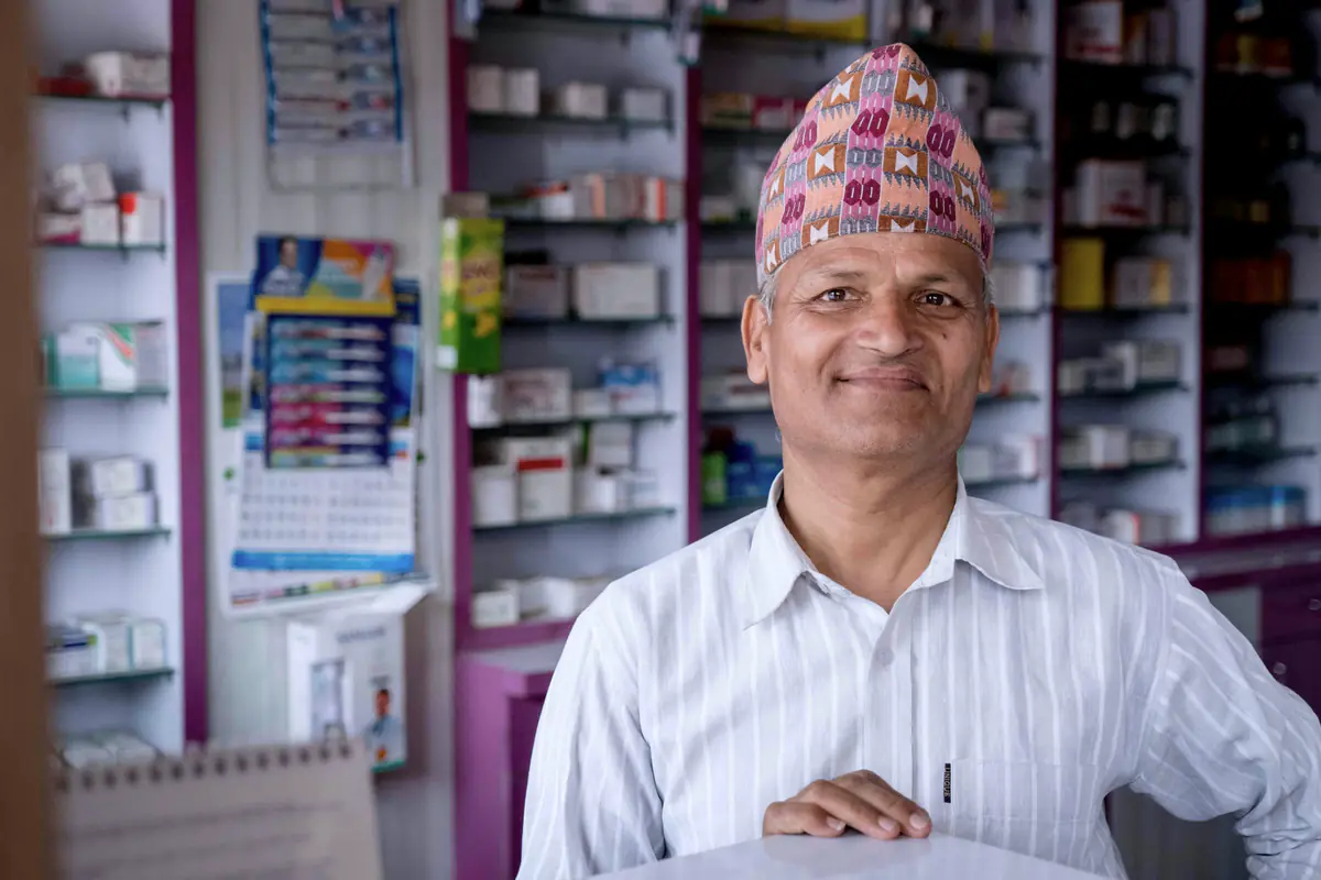A smiling pharmacist at the counter of a pharmacy lined with medicine shelves