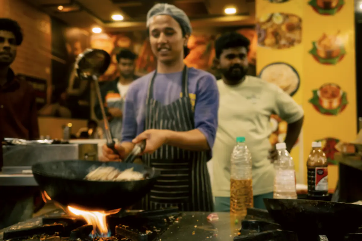 A cook tossing food in a wok over an open flame in a busy restaurant kitchen